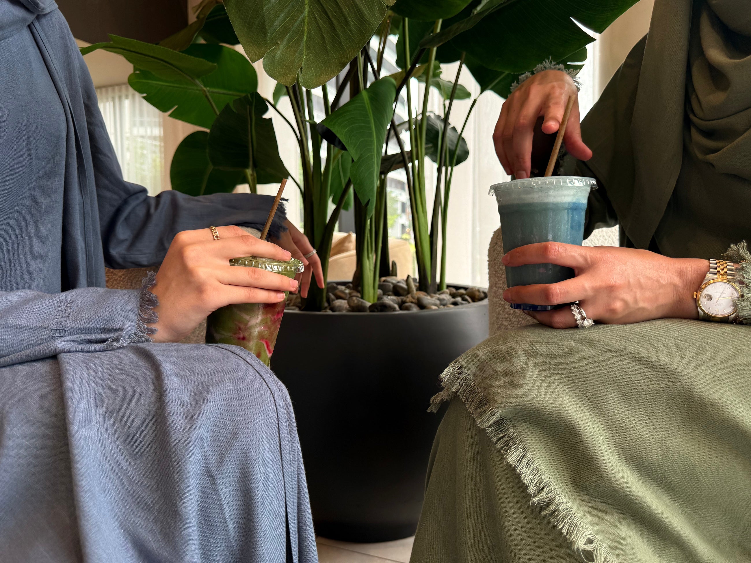 Two women in HAYAR linen abayas, Petroleum and Sage, sharing iced drinks